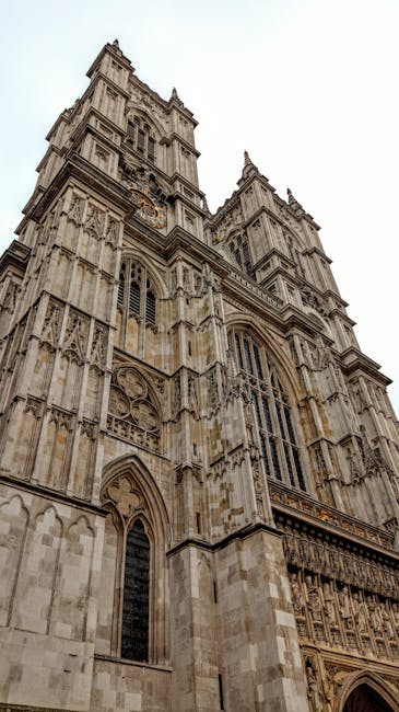 A wide-angle view of Westminster Abbey's Gothic architecture featuring two tall, ornate towers with pointed spires and a large rose window, situated behind a stone column topped with a detailed statue